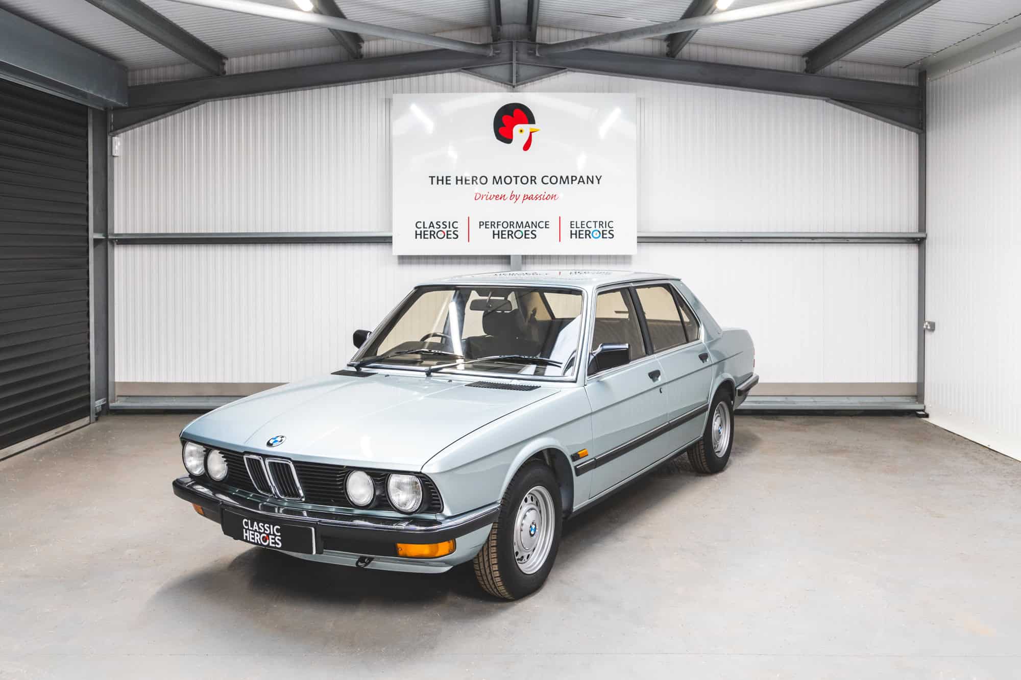 Front quarter view of a sapphire blue BMW E28 525 Eta with chrome bumpers and steel wheels under a Hero Motor Company sign.