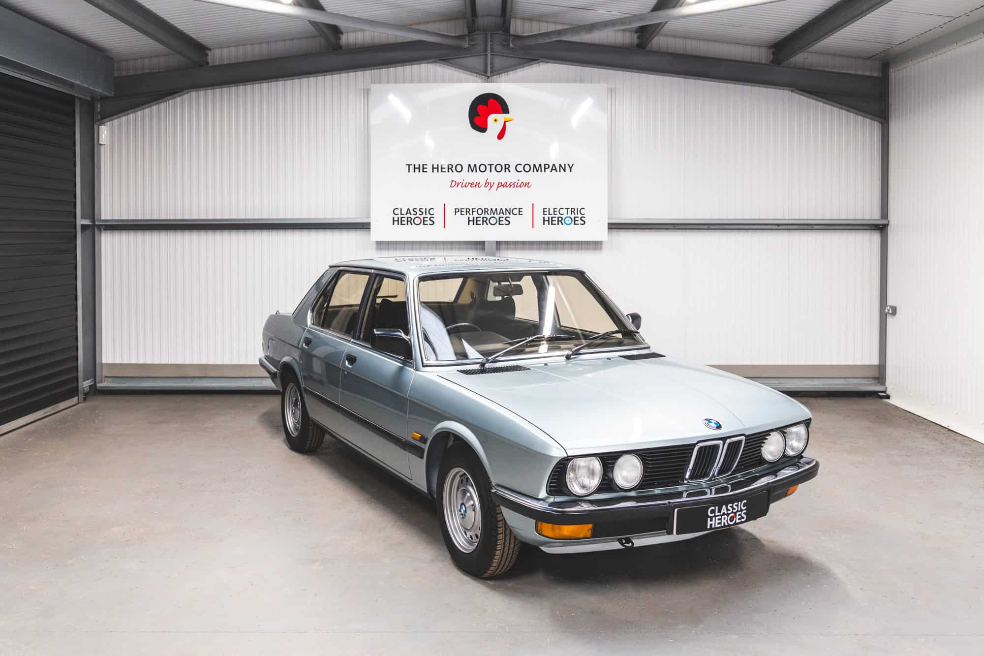 Offside front quarter of BMW E28 525 Eta in classic sapphire blue under a Hero Motor Company Sign.