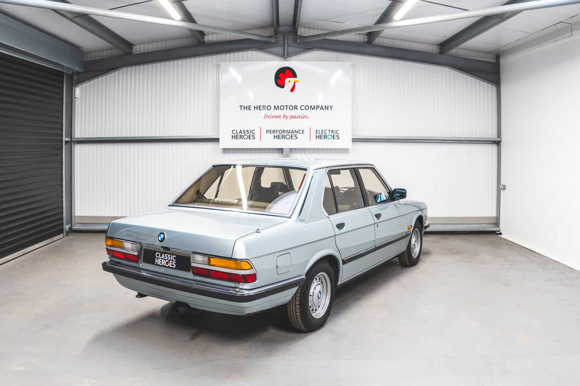 Drivers side rear quarter view of a BMW E28 525 Eta in sapphire blue with chrome bumpers under a Hero Motor Company sign.