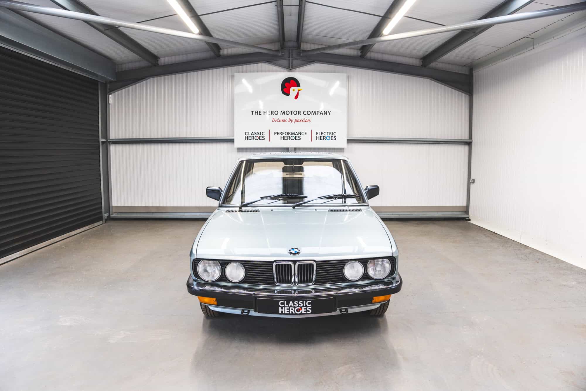 Front end view of an E28 BMW 525 Eta with chrome bumpers and sapphire blue paint under a Hero Motor Company sign.