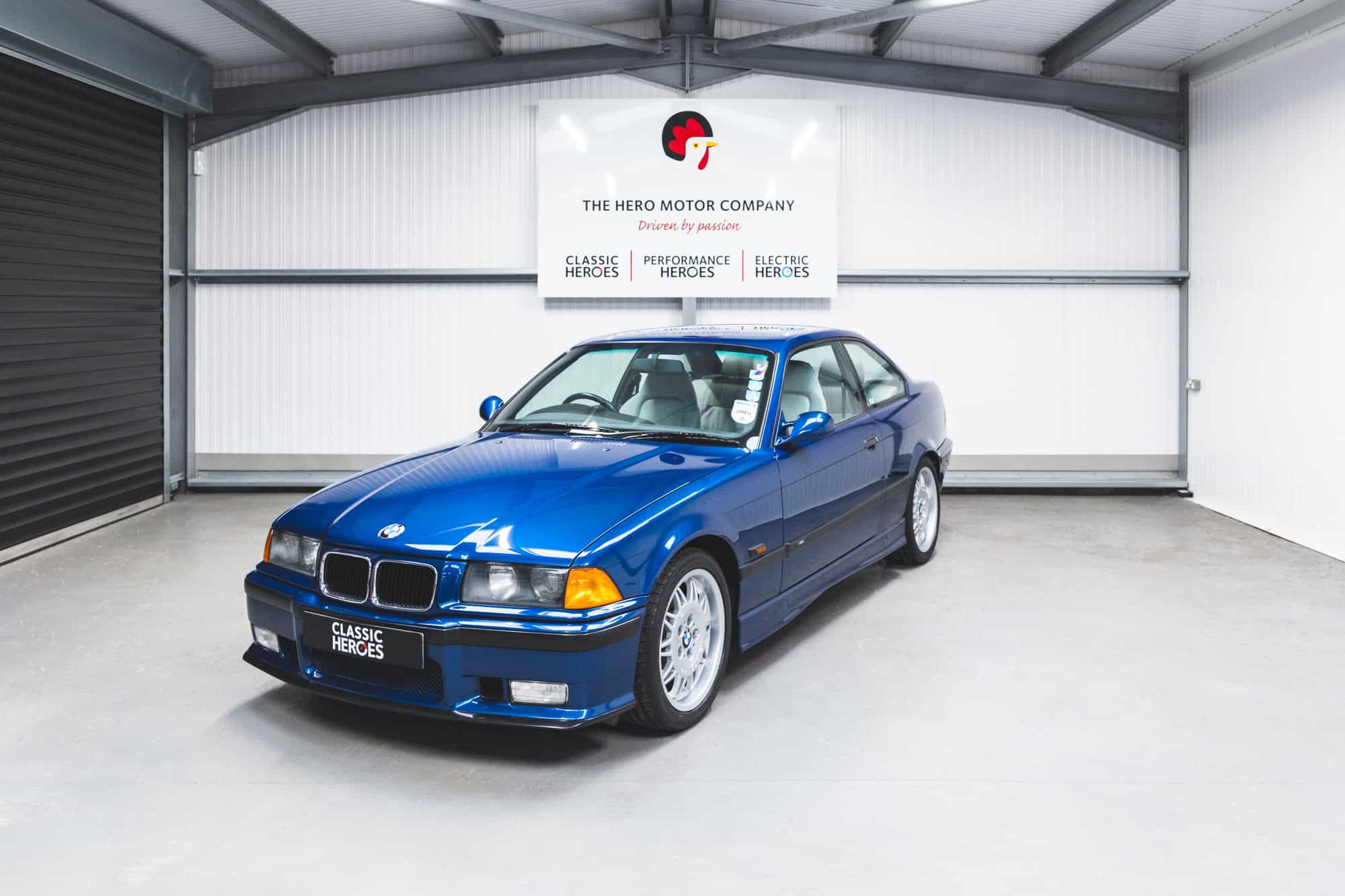 Front quarter view of a BMW E36 M3 Coupe in Avus blue with light Hell grey leather interior visible through a tinted windscreen.