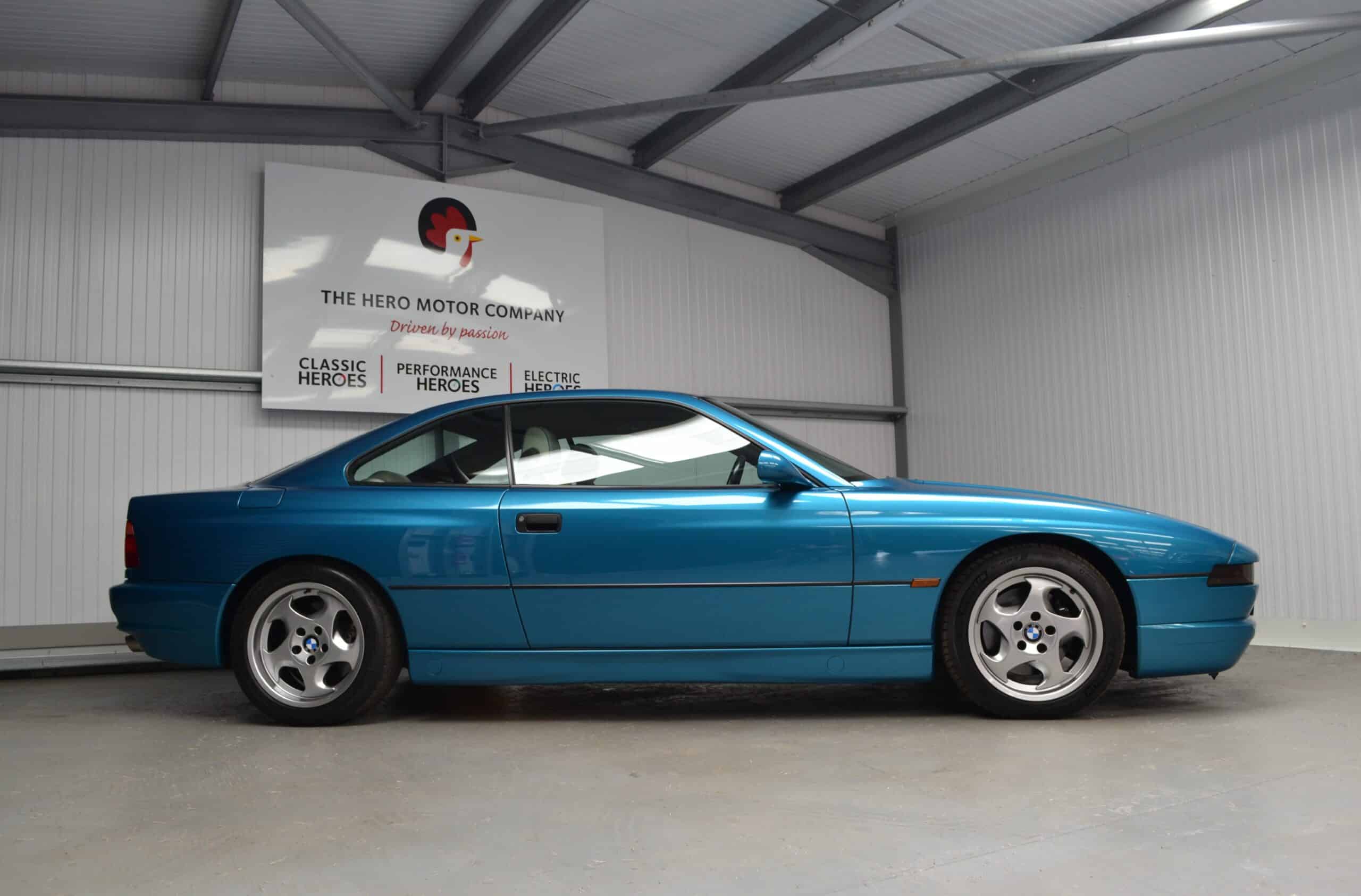 Side profile of a BMW E31 840ci in Atlantis Blue under a Hero Motor Company sign.