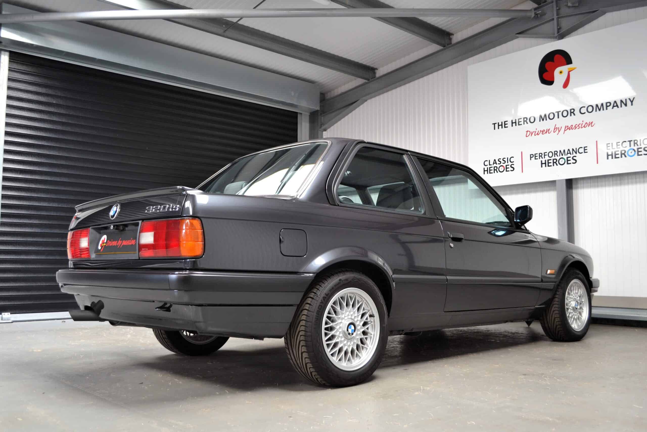 BMW E30 320is Diamond Black offside rear quarter under a Hero Motor Company sign.