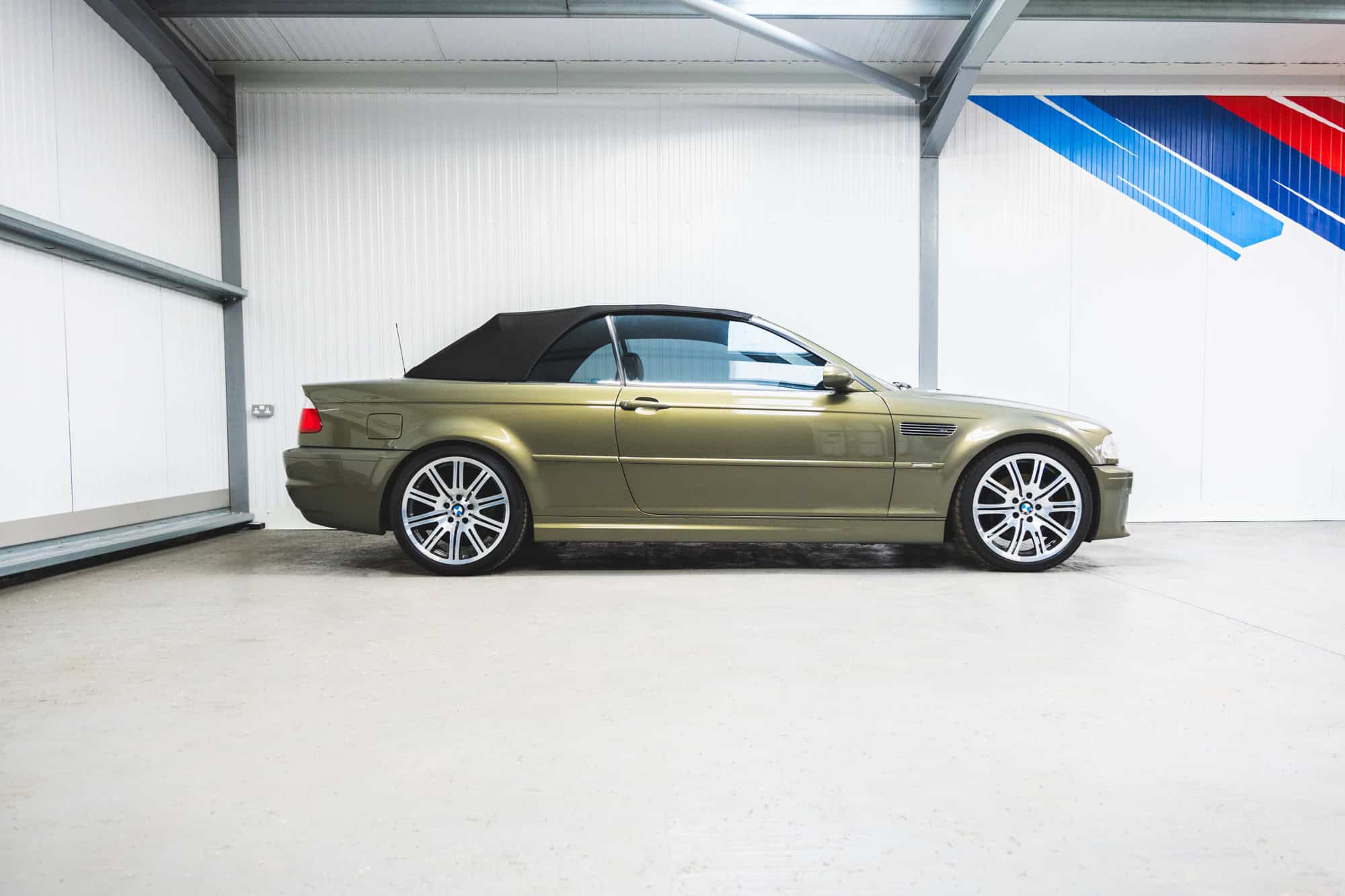 Side view of a BMW E46 M3 Convertible with the roof up and alloy wheels and Messing Metallic paint against an M motorsport wall.