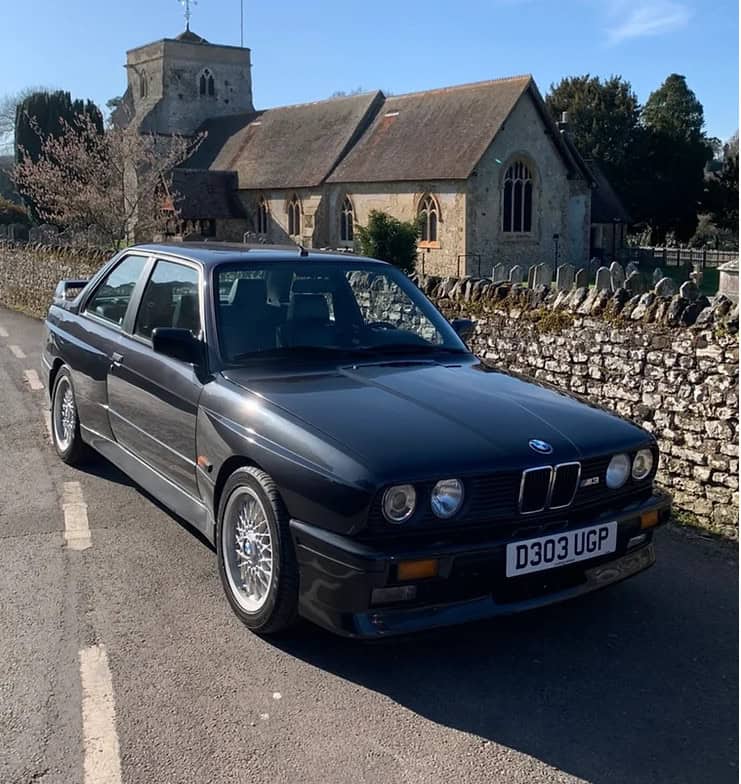 Offside exterior of black M3 parked by a country wall.