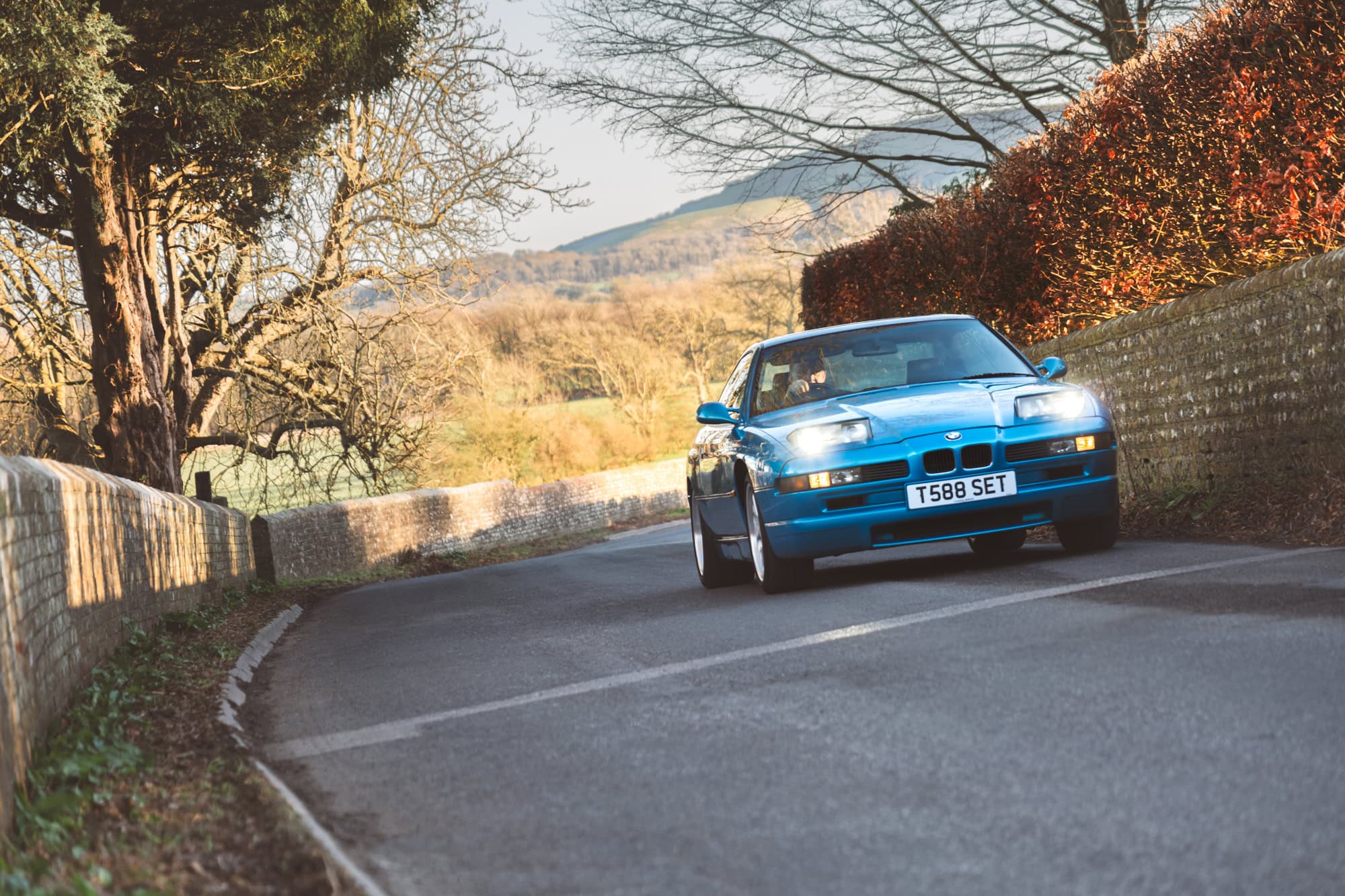 A blue BMW E31 850 CSI driving up a country road and going very fast.