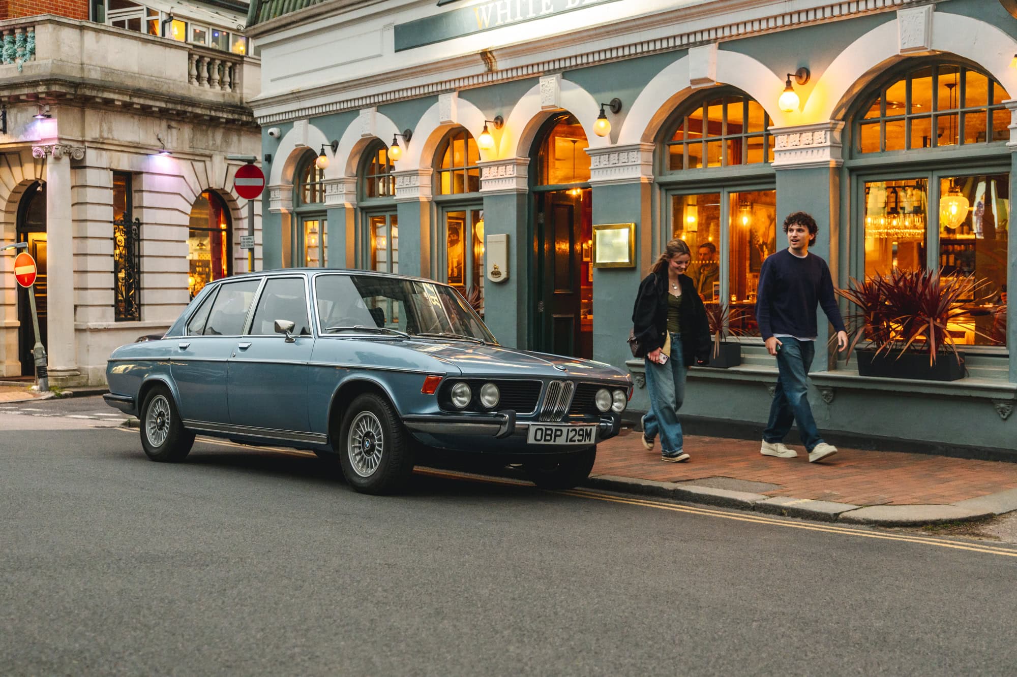 A BMW E3 3.0 Si Saloon in Fjord Blue is parked outside a trendy wine bar in the Pantiles Tunbridge Wells as a young couple walk by.