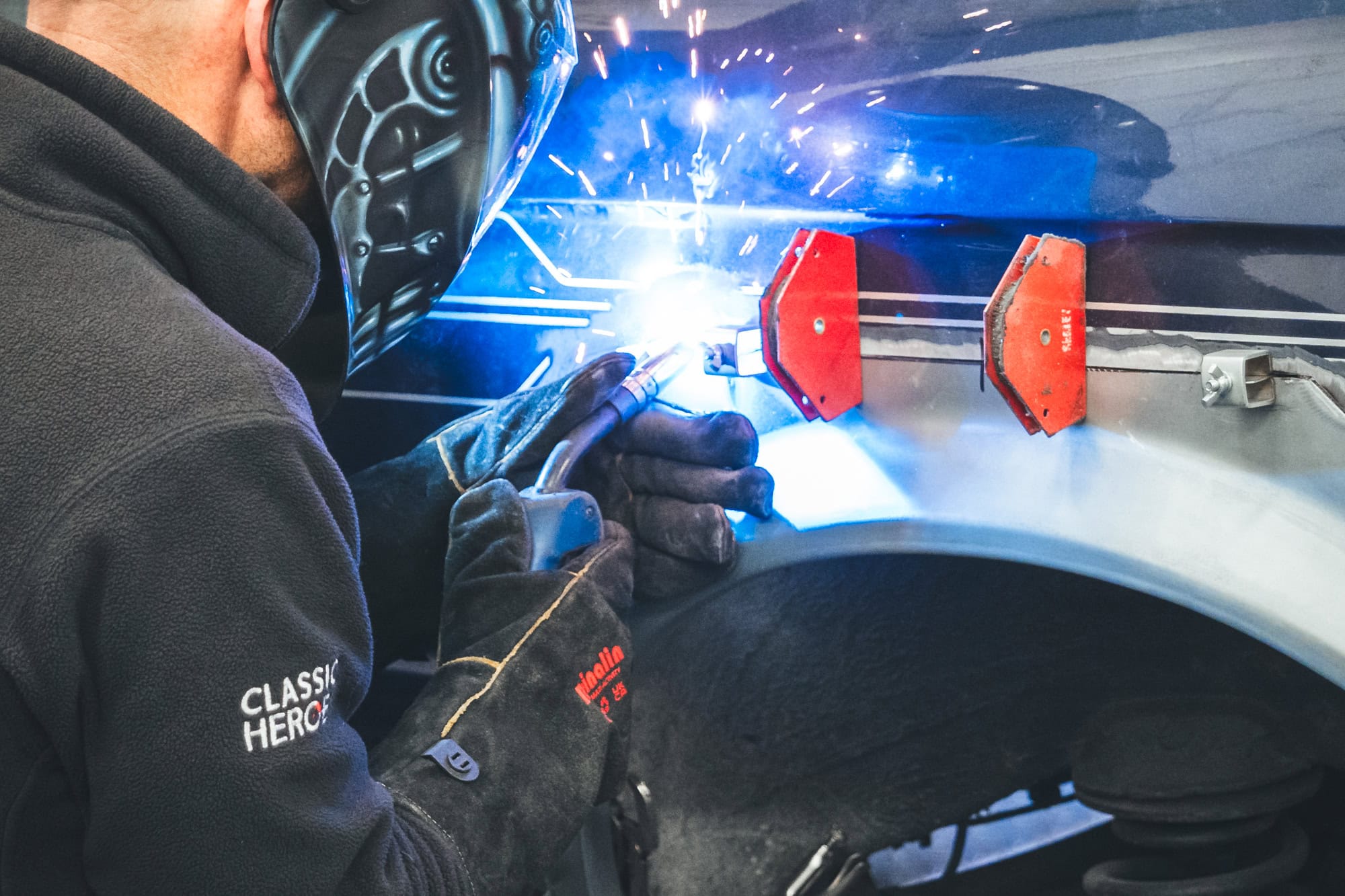 A skilled mechanic and fabricator welding a new arch repair onto a BMW E30 Alpina C2 wearing a hero motor company uniform mobile image.