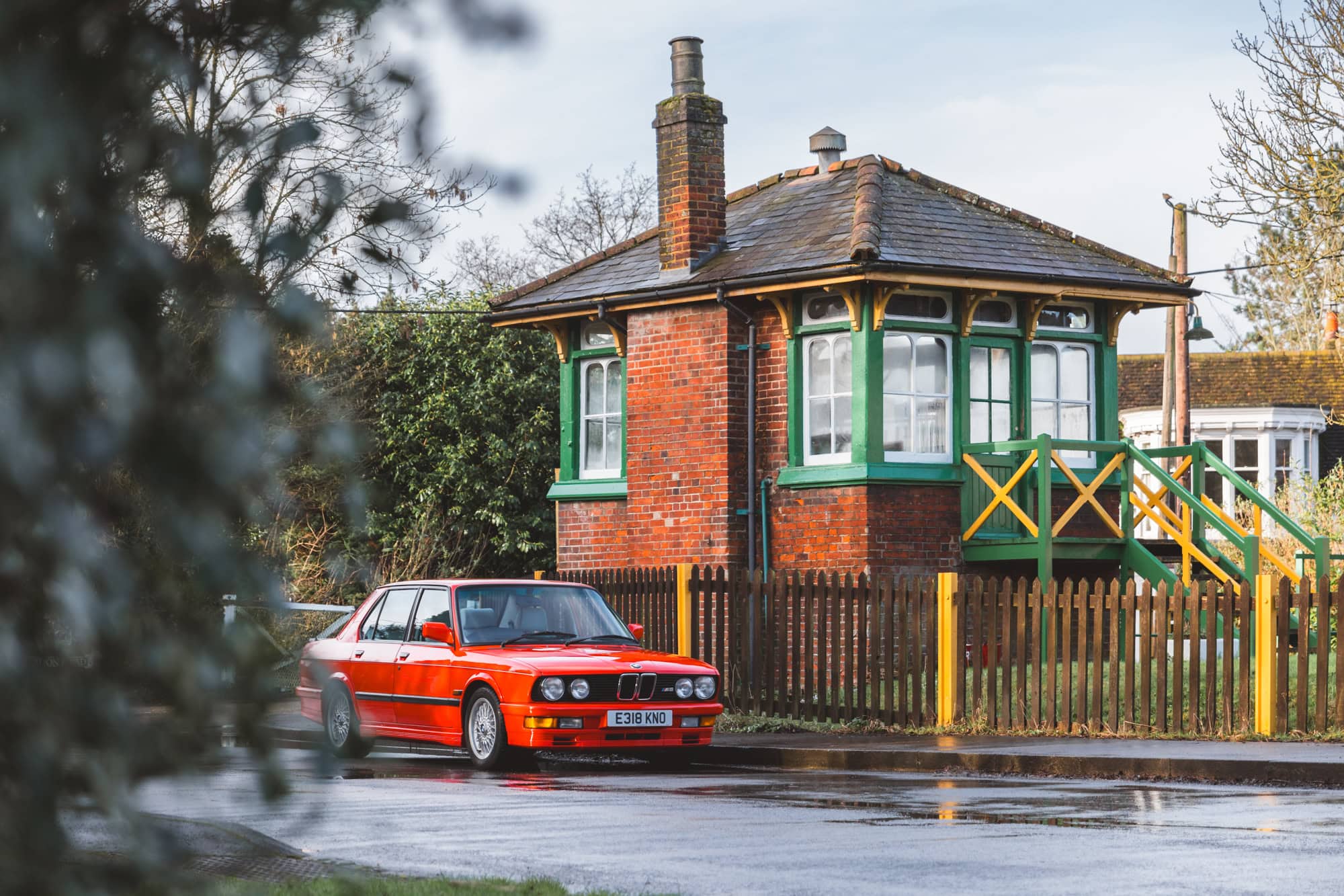 An orange BMW E28 M5 parked in a rural village next to an old train station signal box.