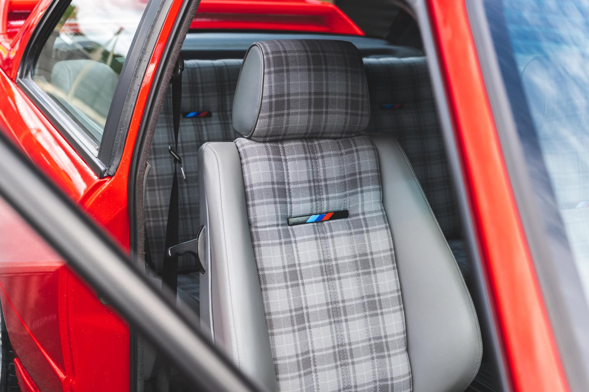 A red BMW E30 M3 front seat in grey chequered upholstery.