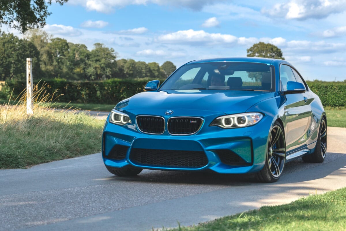 A metallic blue BMW M2 drives around a rural country road.