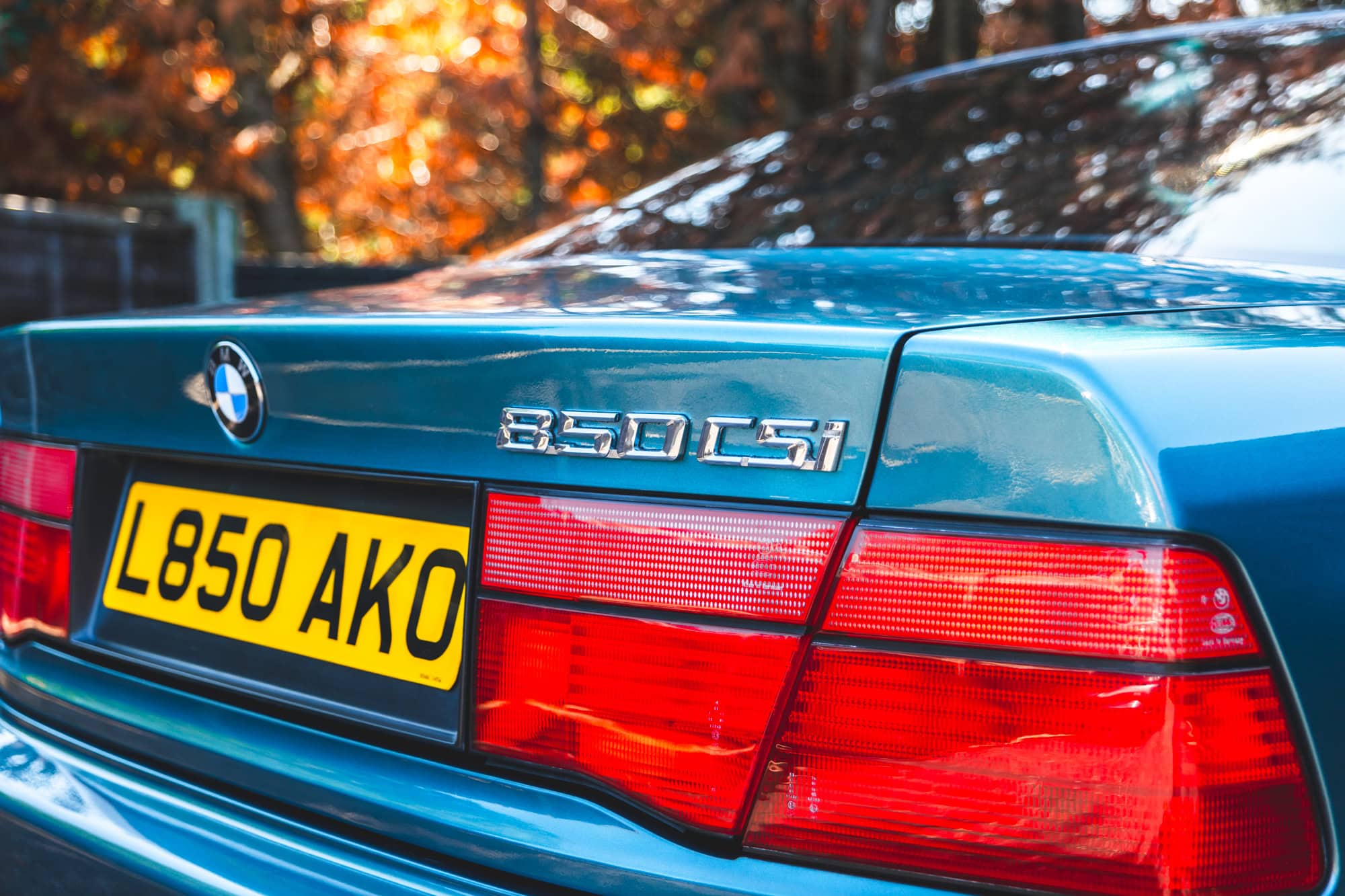 Rear quarter and tail lights of a BMW E31 850csi in Barbados green with orange autumn leaves in the background.