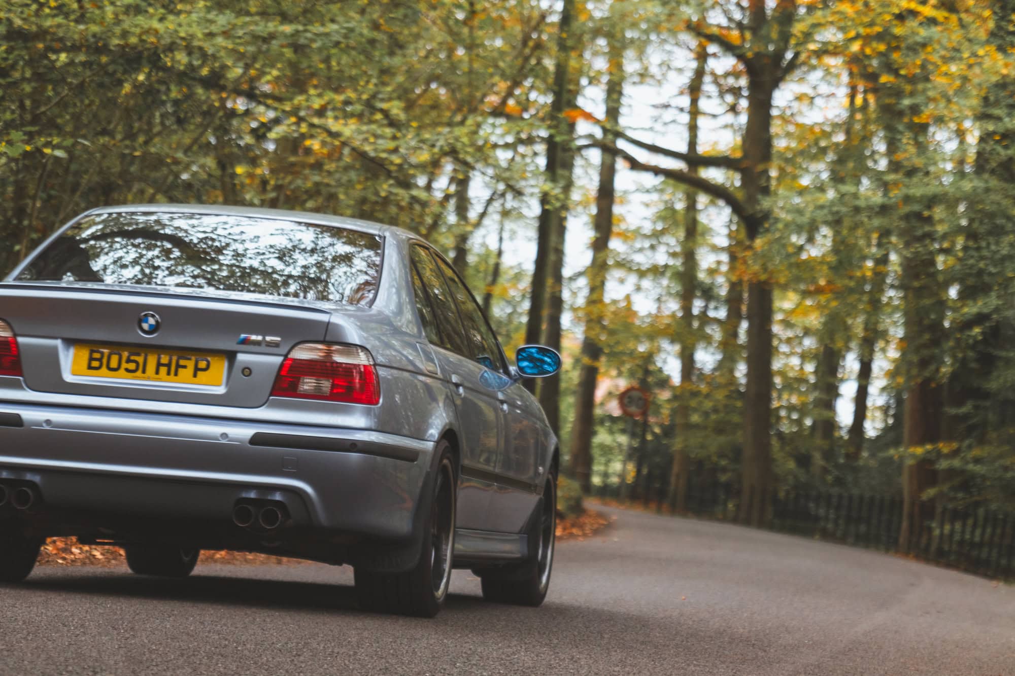 A Silverstone blue BMW E39 M5 drives dynamically down a sweeping autumn road.