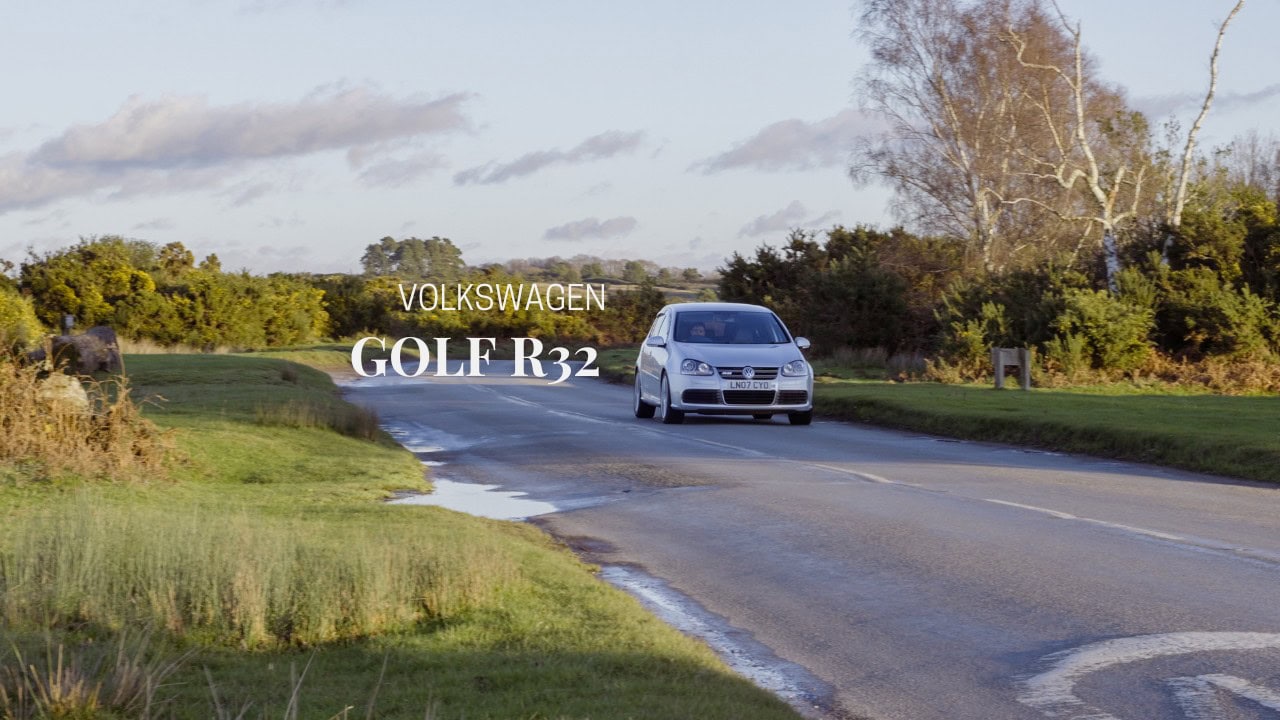 A silver Volkswagen Golf GTI R32 drives down a forest road at sunset.