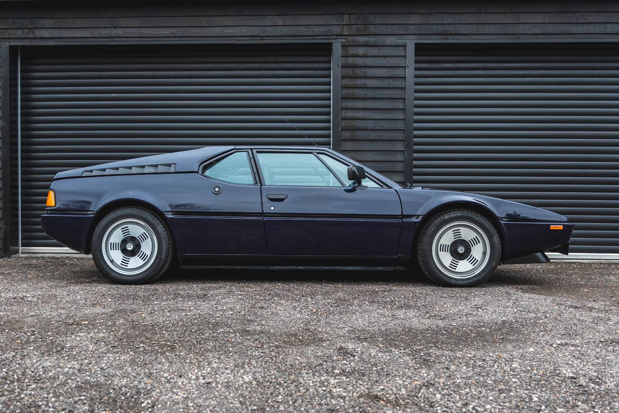 Offside exterior view of 1980 E26 BMW M1 Nachtblau showing alloy wheels and in the background black roller shutters on a timber building