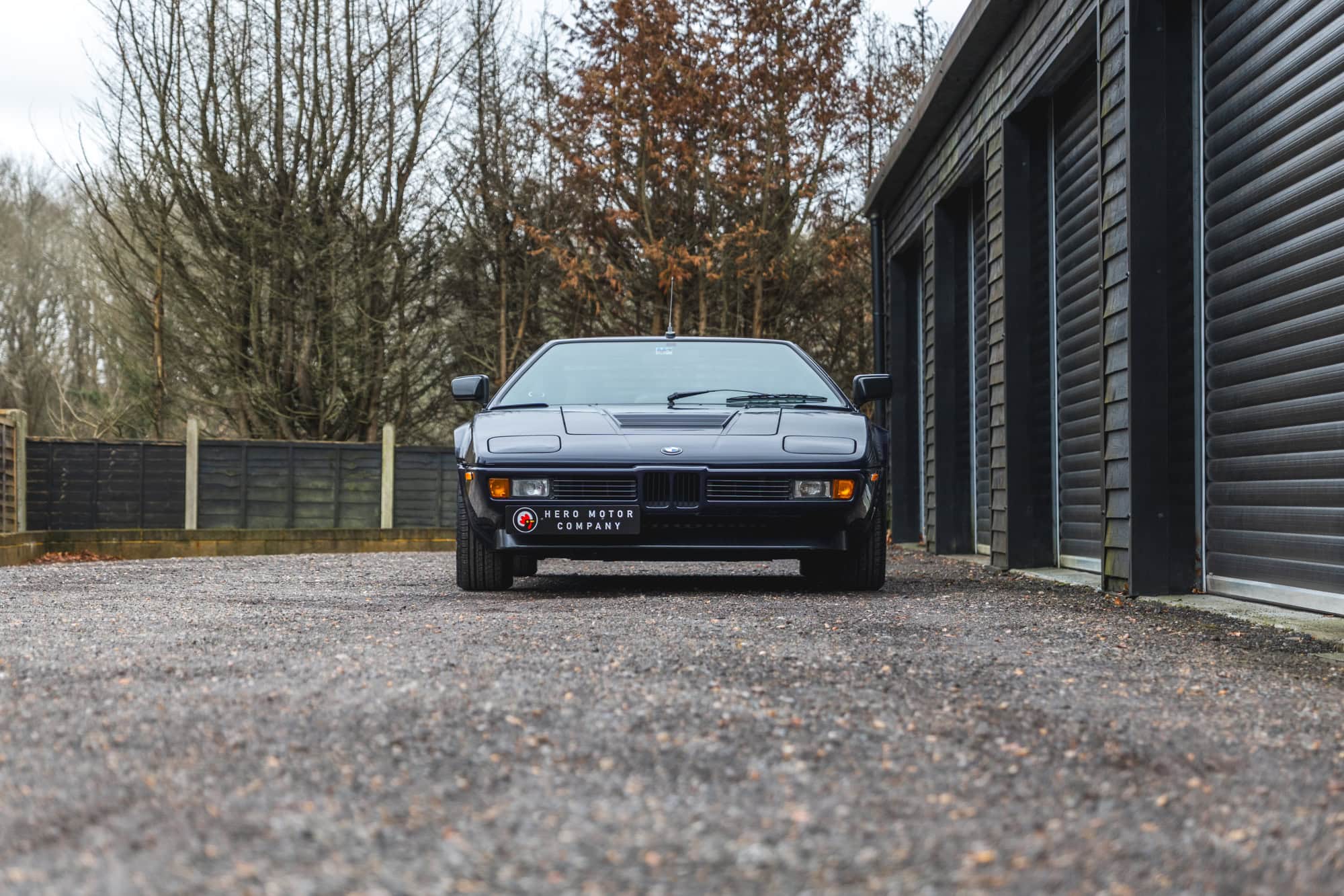 Front exterior view of 1980 E26 BMW M1 Nachtblau with black roller shutters and trees in the background