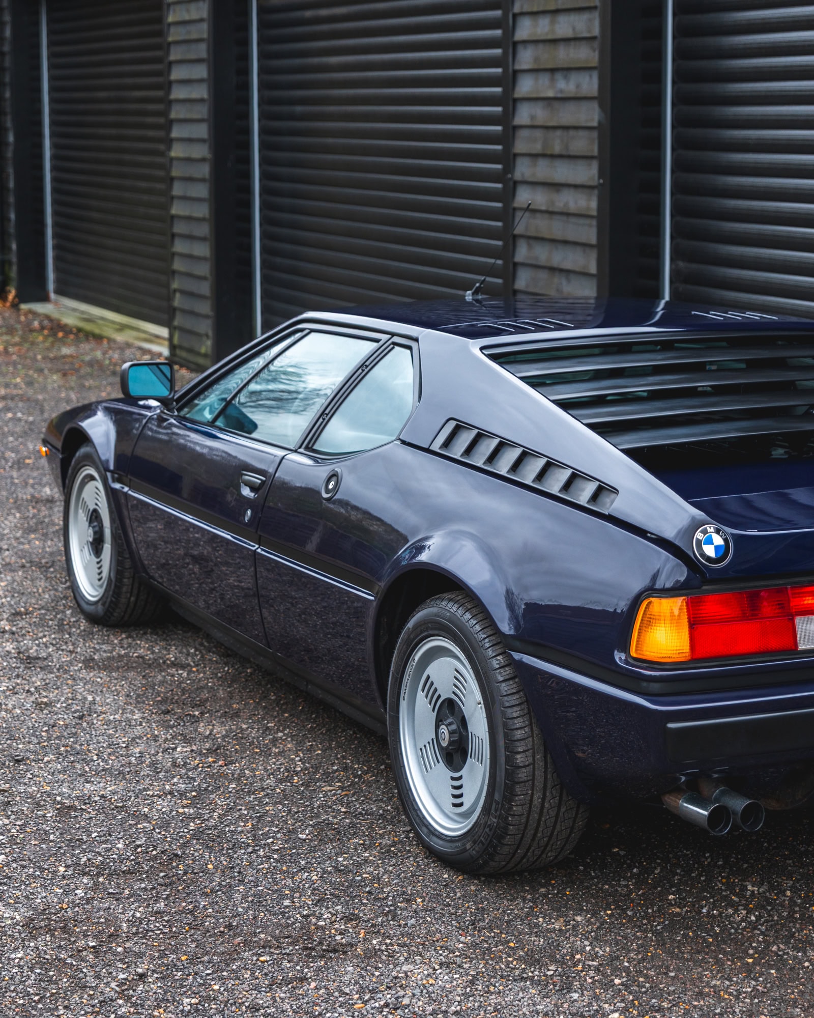 Nearside exterior view of 1980 E26 BMW M1 Nachtblau with black timberclad build with black rollershutters in the background