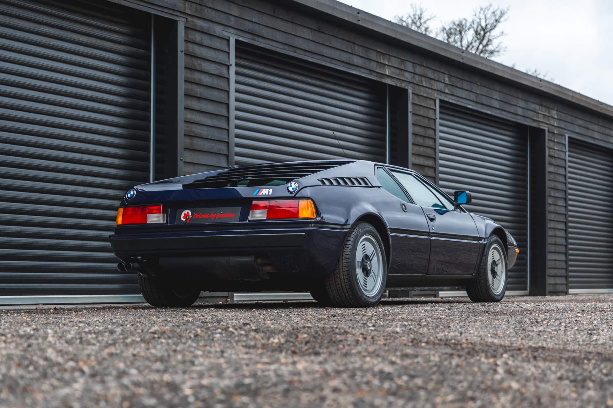 Rear offside exterior of 1980 E26 BMW M1 Nachtblau showing alloy wheels and louvre slats with black timer building in the rear