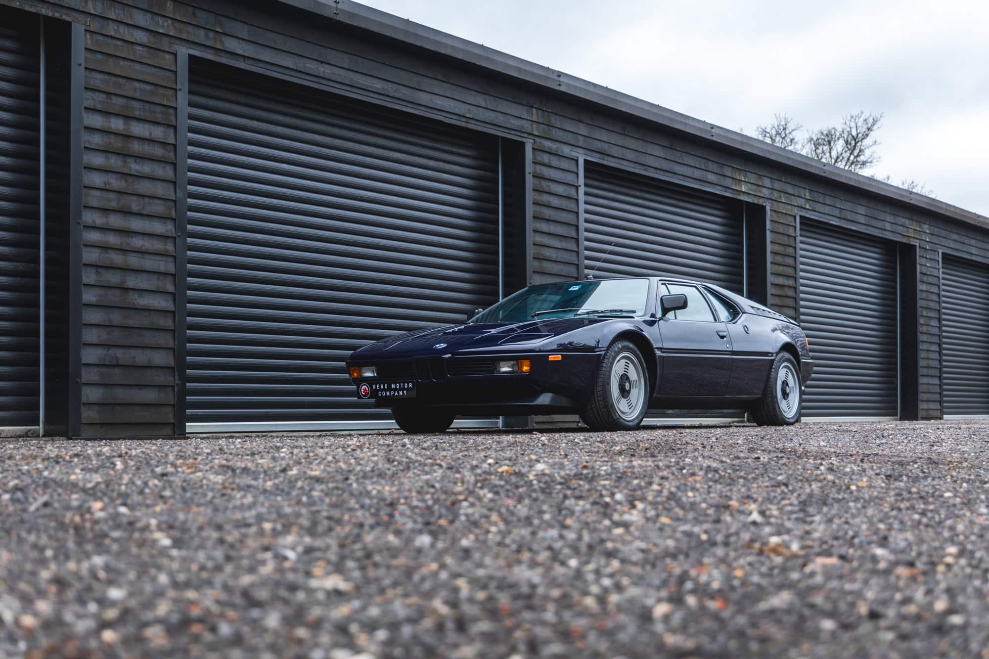 Nearside front exterior of 1980 E26 BMW M1 Nachtblau with black timber building in the background