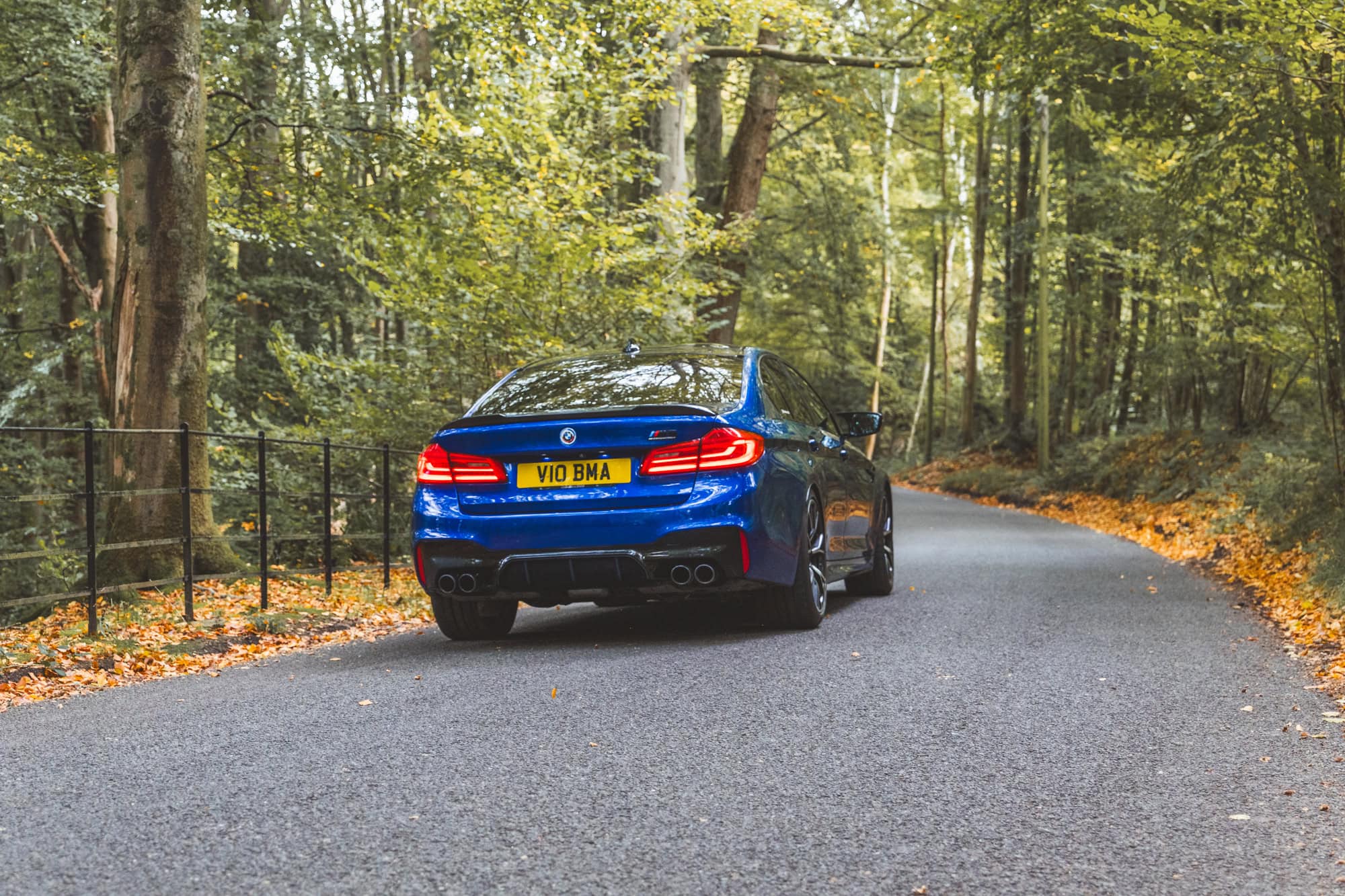 Rear quarter view of a Marina Blue F90 BMW M5 Competition driving away down a forest road.