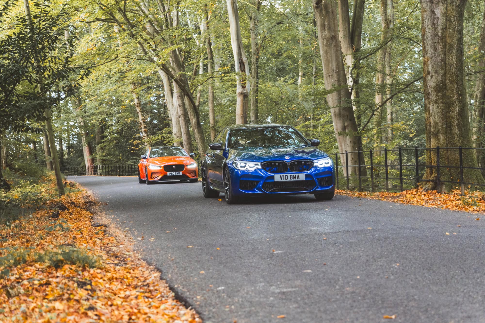 An F90 BMW M5 Competition in Marina Blue being chased down a forest road by a Valencia Orange Project 8 Jaguar.