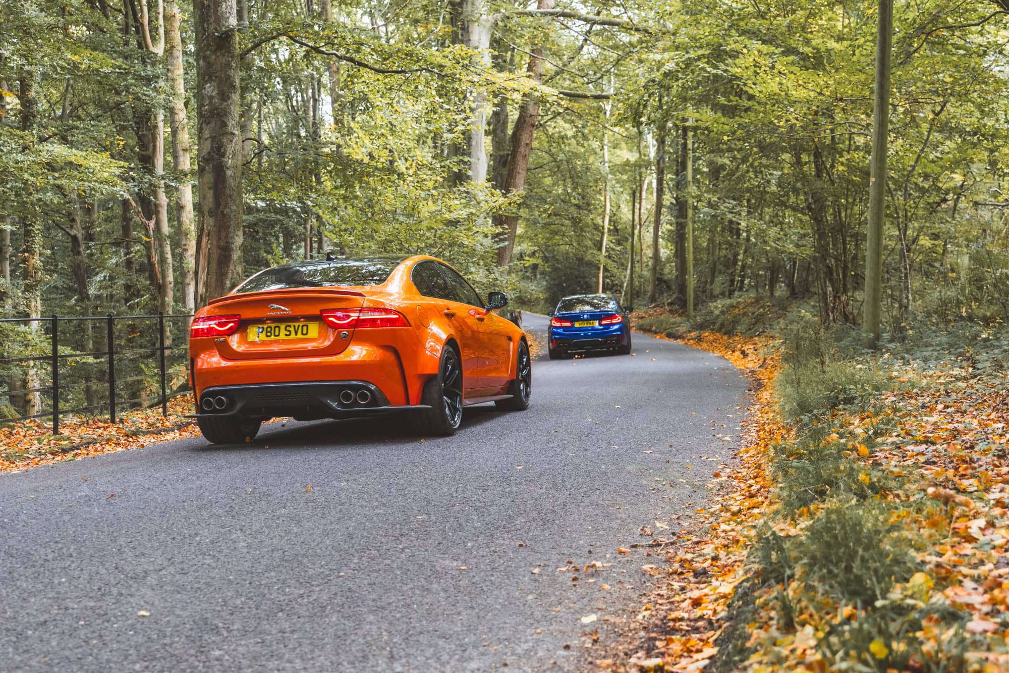 An F90 BMW M5 Competition in Marina Blue being chased down a forest road by a Valencia Orange Project 8 Jaguar.