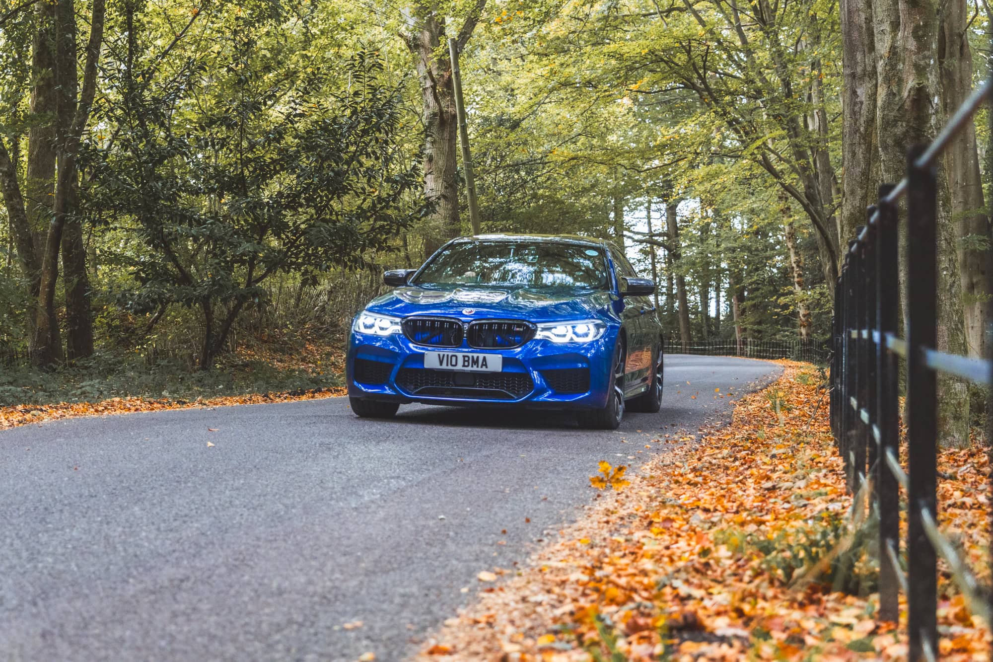 An F90 BMW M5 Competition in Marina Blue driving down a forest road with project gamma air intakes visible.