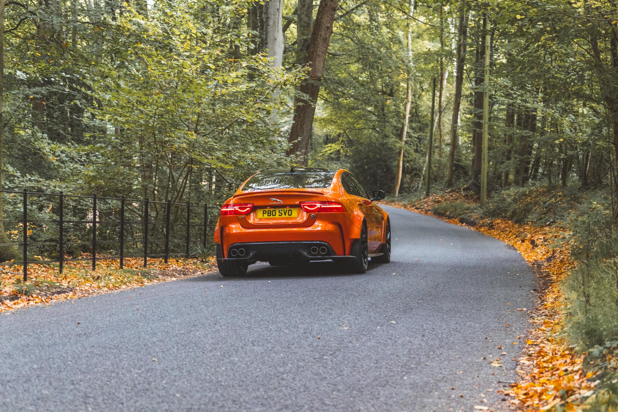 Rear quarter view of a Valencia Orange Jaguar Project 8 saloon driving away down a forest road.