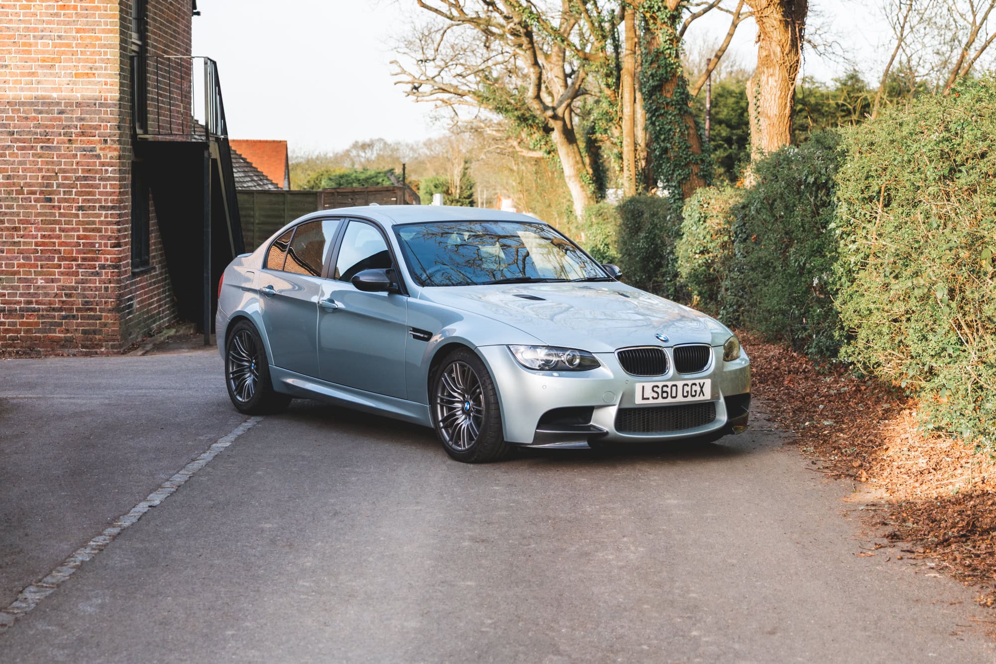 Front offside view of BMW E90 M3 Silverstone II showing alloy wheels parked on a lane with trees a building and fencing in the background
