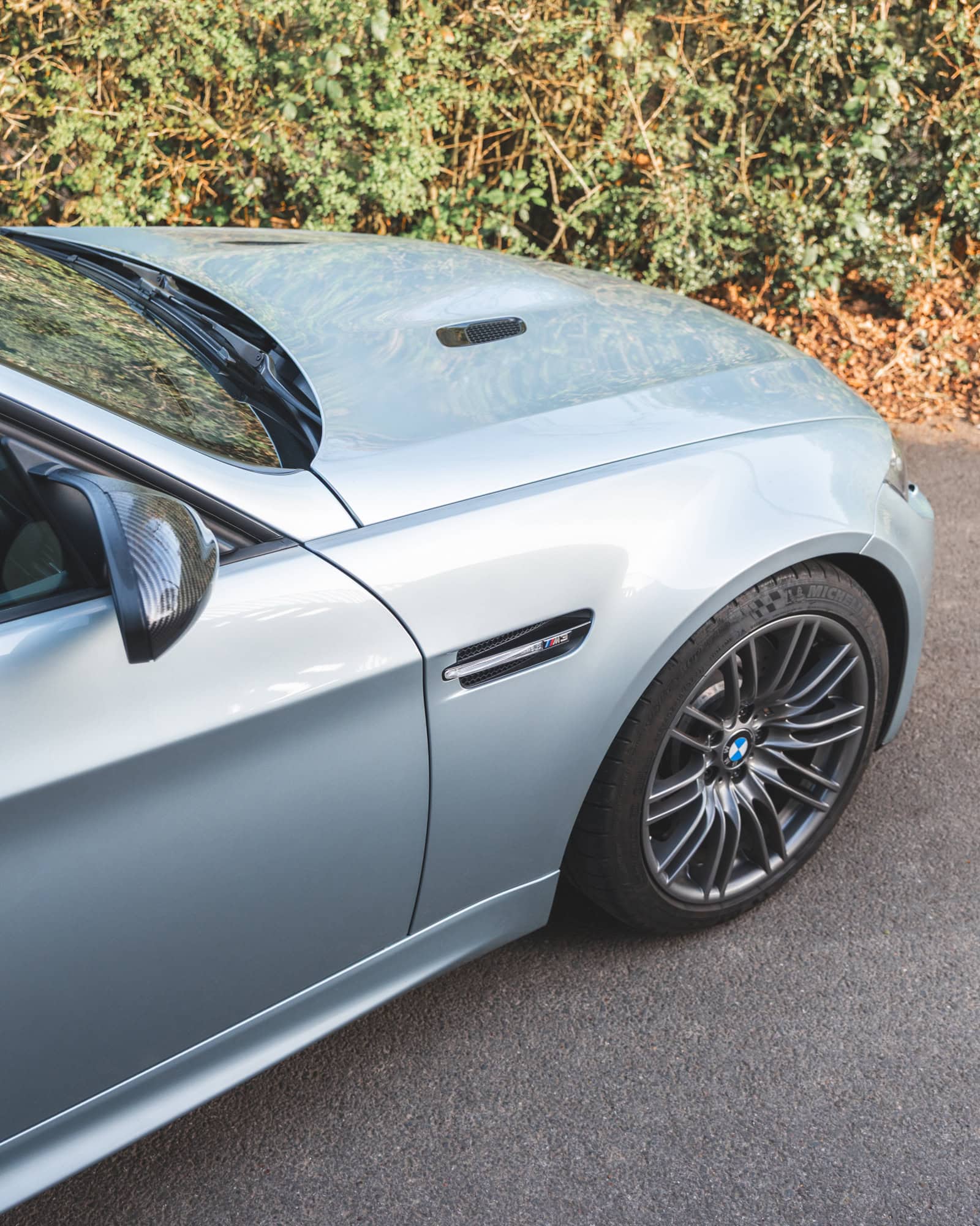 Front quarter of BMW E90 M3 in Silverstone II showing alloy wheel in front of a hedge and brown leaves