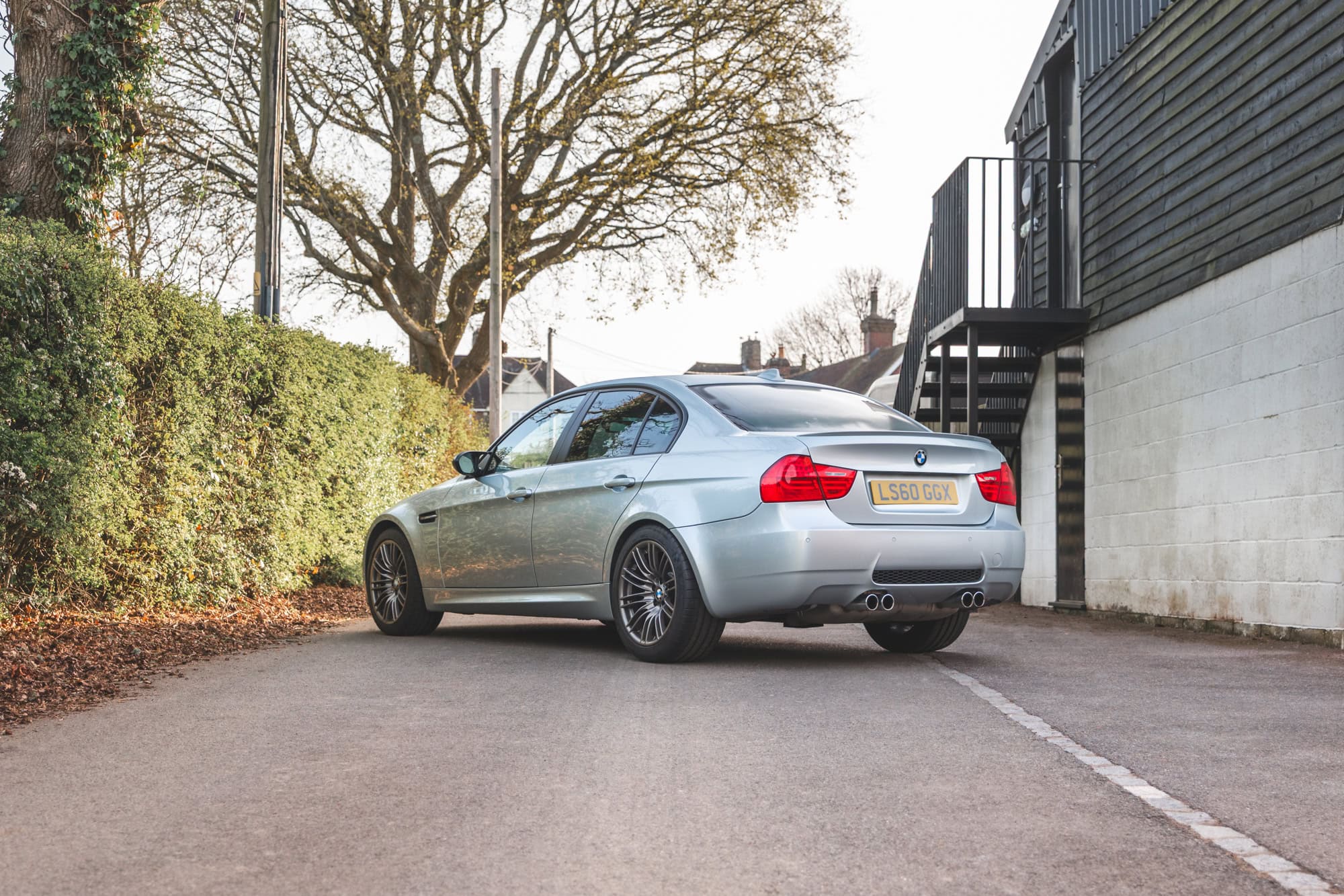 Rear nearside exterior of BMW E90 M3 Silverstone II parked on a lane with hedge on nearside and building with black steps on the offside