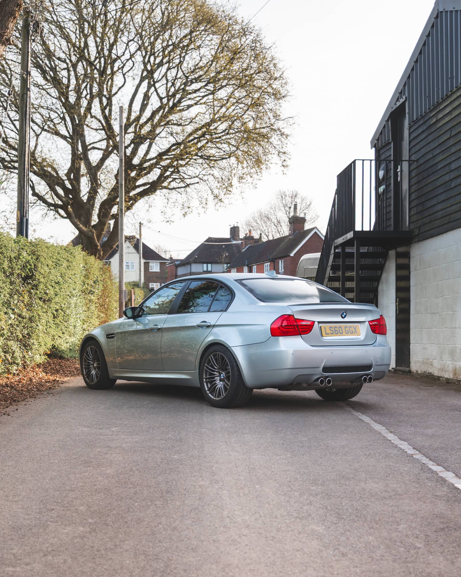 Rear nearside exterior of BMW E90 M3 Silverstone II parked on a lane with hedge on nearside and building with black steps on the offside