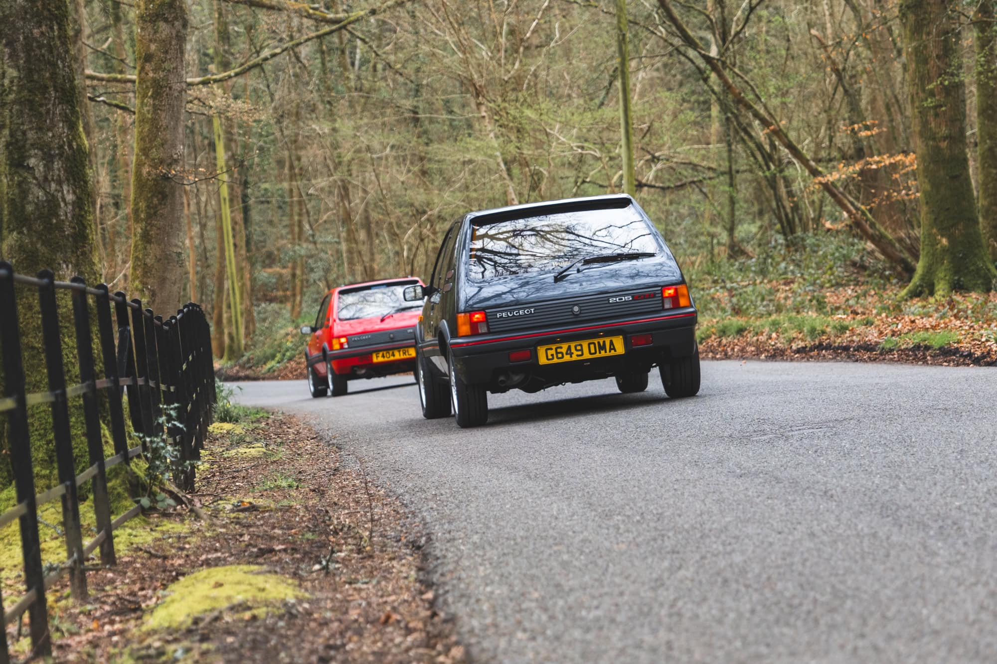 Rear view of two Peugeot 205 GTI on a country lane with trees in the back ground In gris granite and cherry red