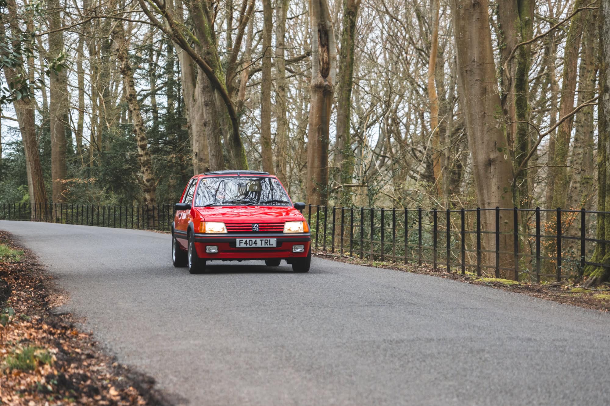 Front view of cherry red Peugeot 205 GTI on a country road showing headlamps and front grill with metal fencing and trees in the background