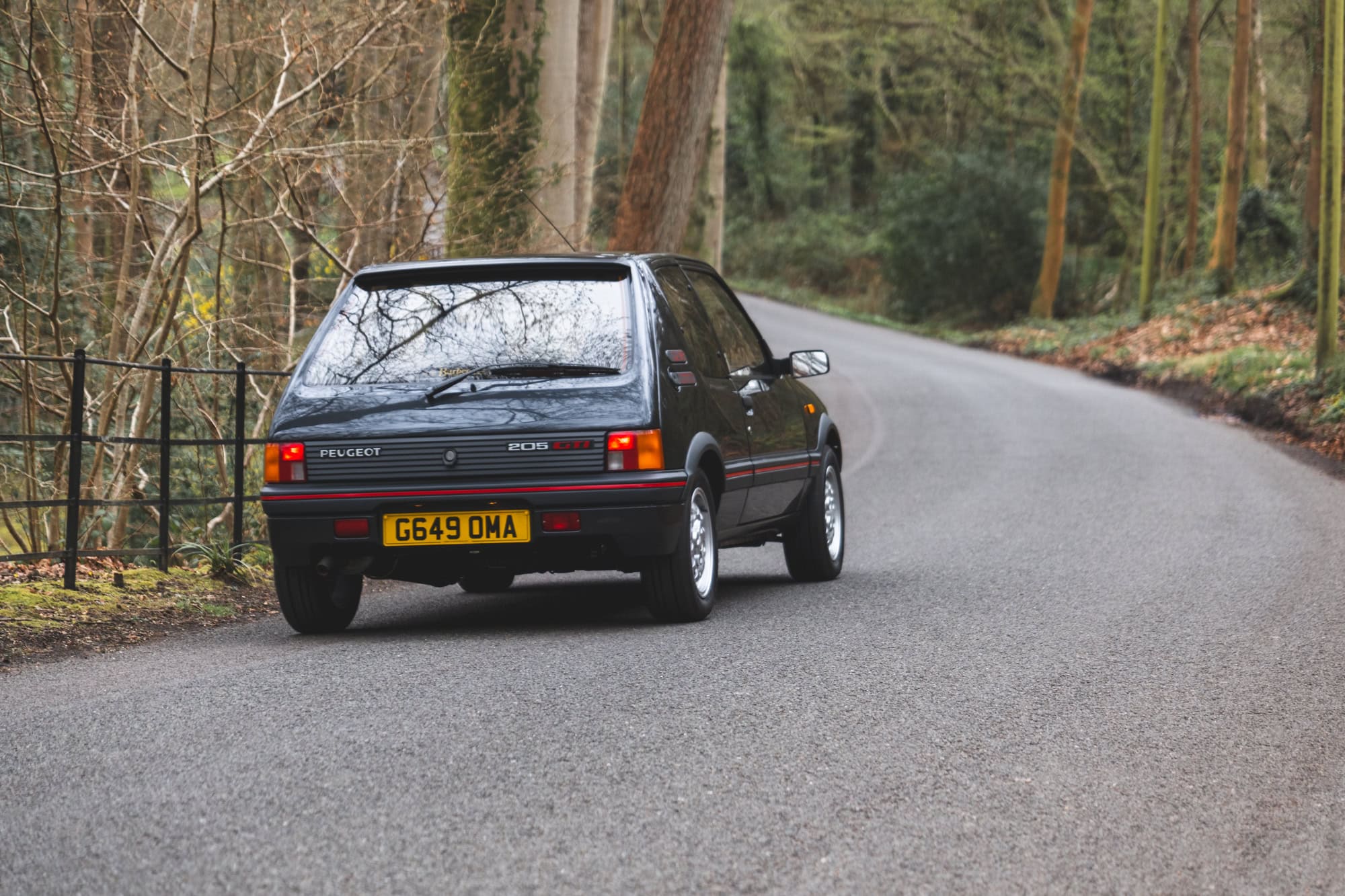 Rear view of gris granite Peugeot 205 GTI on a country lane with fencing and trees in the foreground