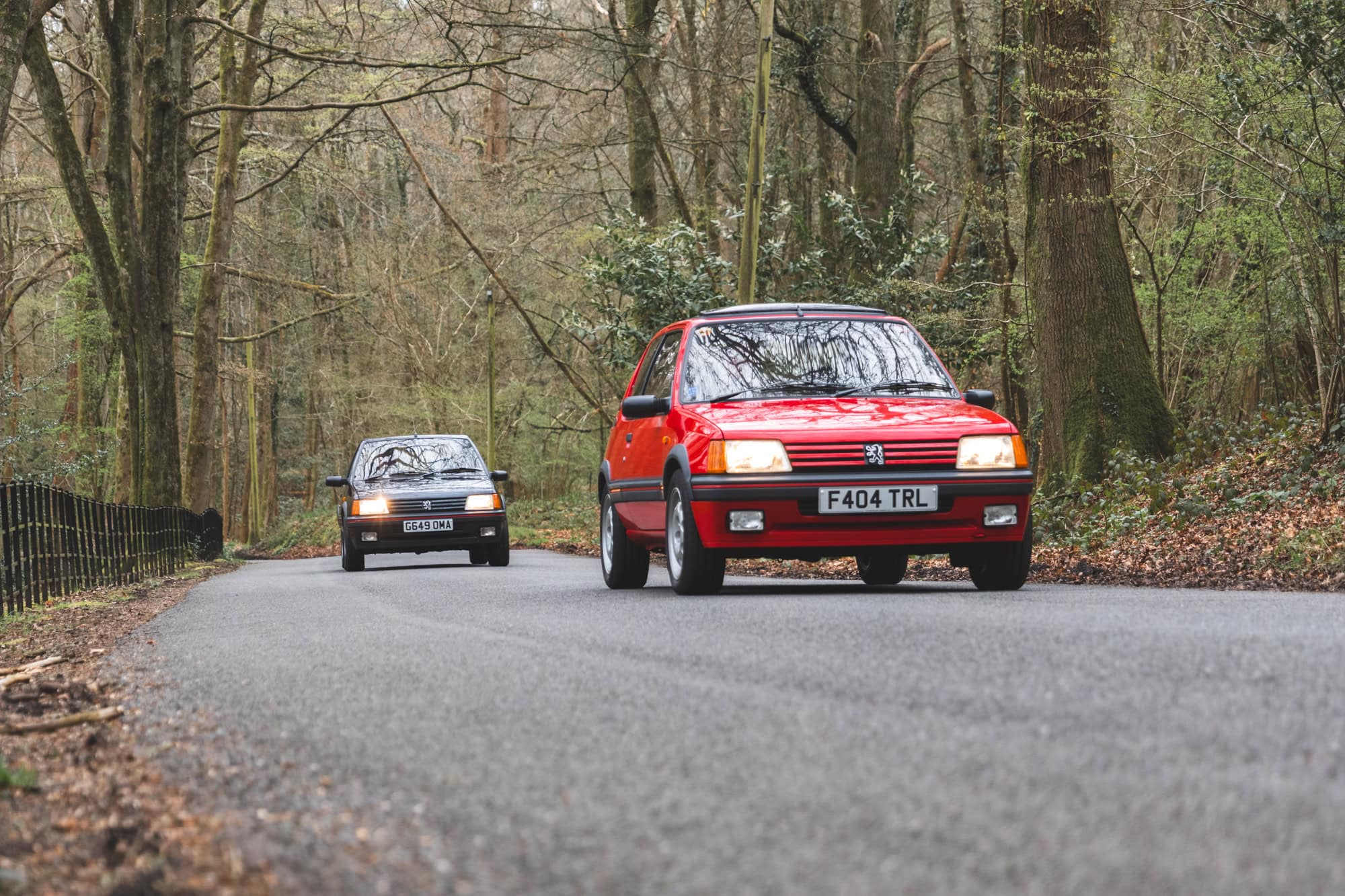 Cherry red Peugeot 205 GTI driving in front of the gris granite 205 GTI on a country lane with trees in the background and brown leaves on the verge