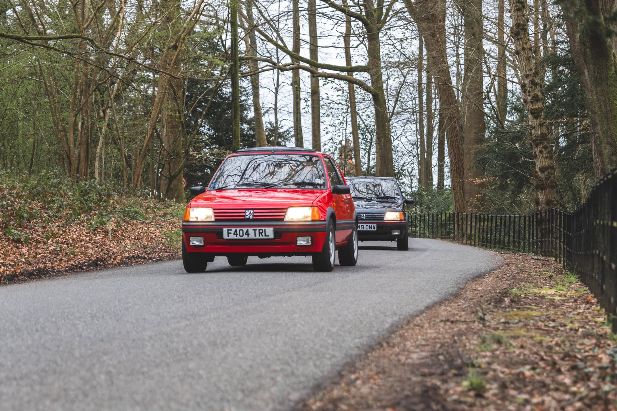 Two Peugeot 205 GTI on a country lane cherry red in front with gris granite at the rear brown leaves at the road side with trees in the background