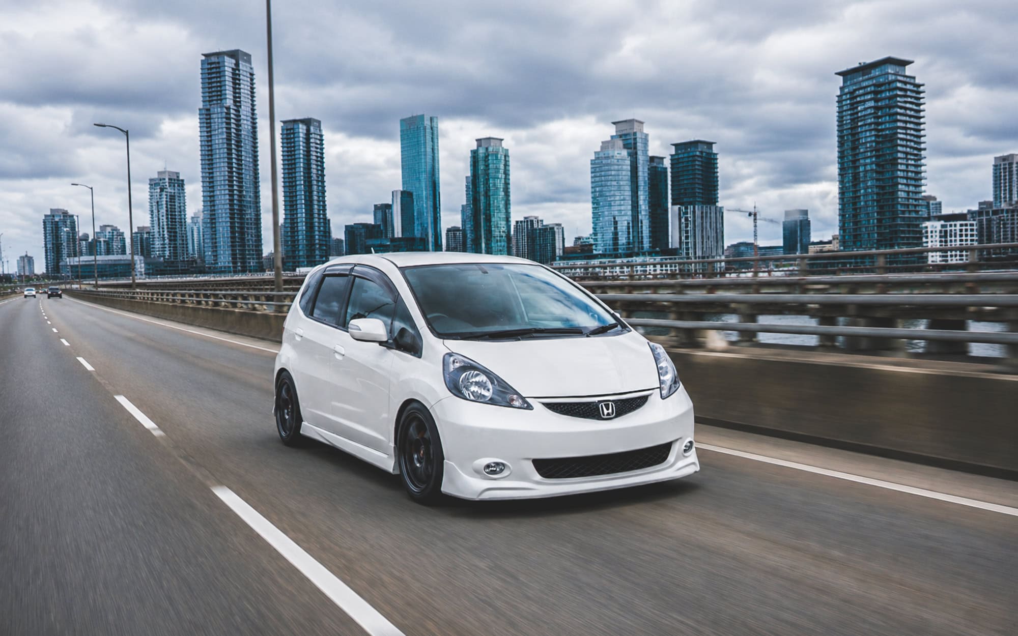 A white Honda Jazz driving in an urban highway.